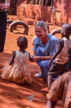 Eine Mitarbeiterin kniet im roten Sand und reicht einem kleinen Mädchen die Hand.