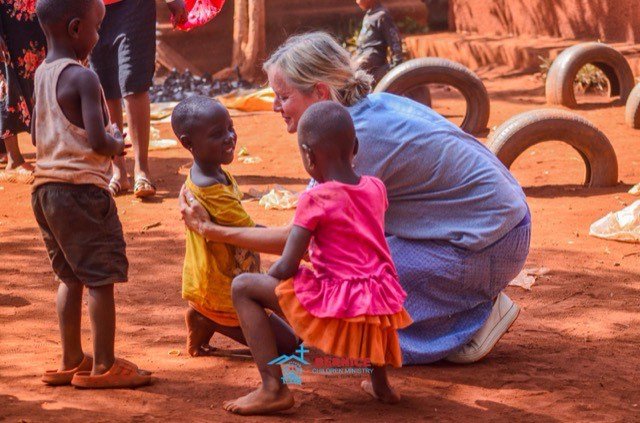 Eine Mitarbeiterin kniet auf dem Sandplatz und spielt mit drei Kindern.