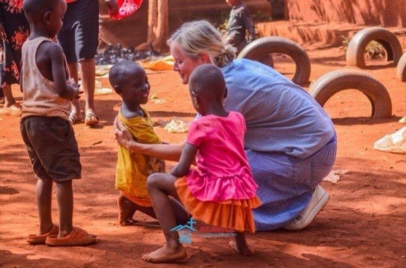 Eine Mitarbeiterin kniet auf dem Sandplatz und spielt mit drei Kindern.