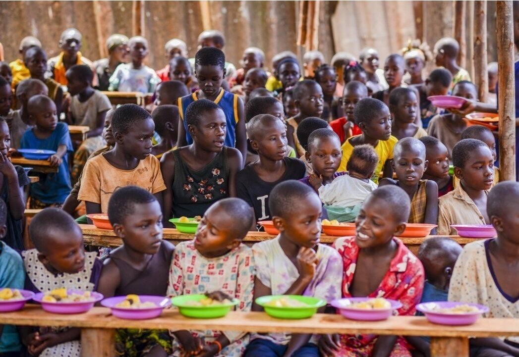 Viele Kinder sitzen an langen Bänken, vor ihnen bunte Teller mit Essen.